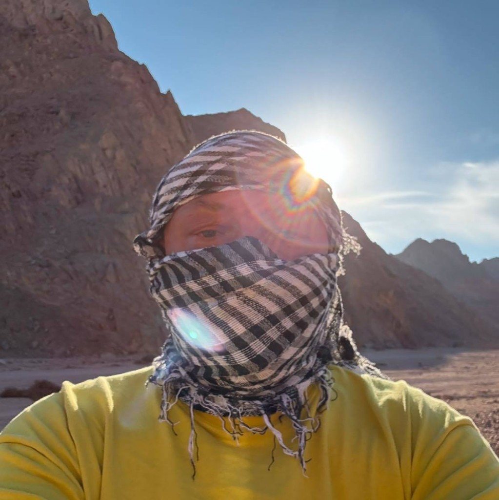 Alex (DC1AS) mit Keffiyeh und gelbem Shirt in der ägyptischen Wüste vor Felskulisse, gegenlicht durch die Mittagssonne